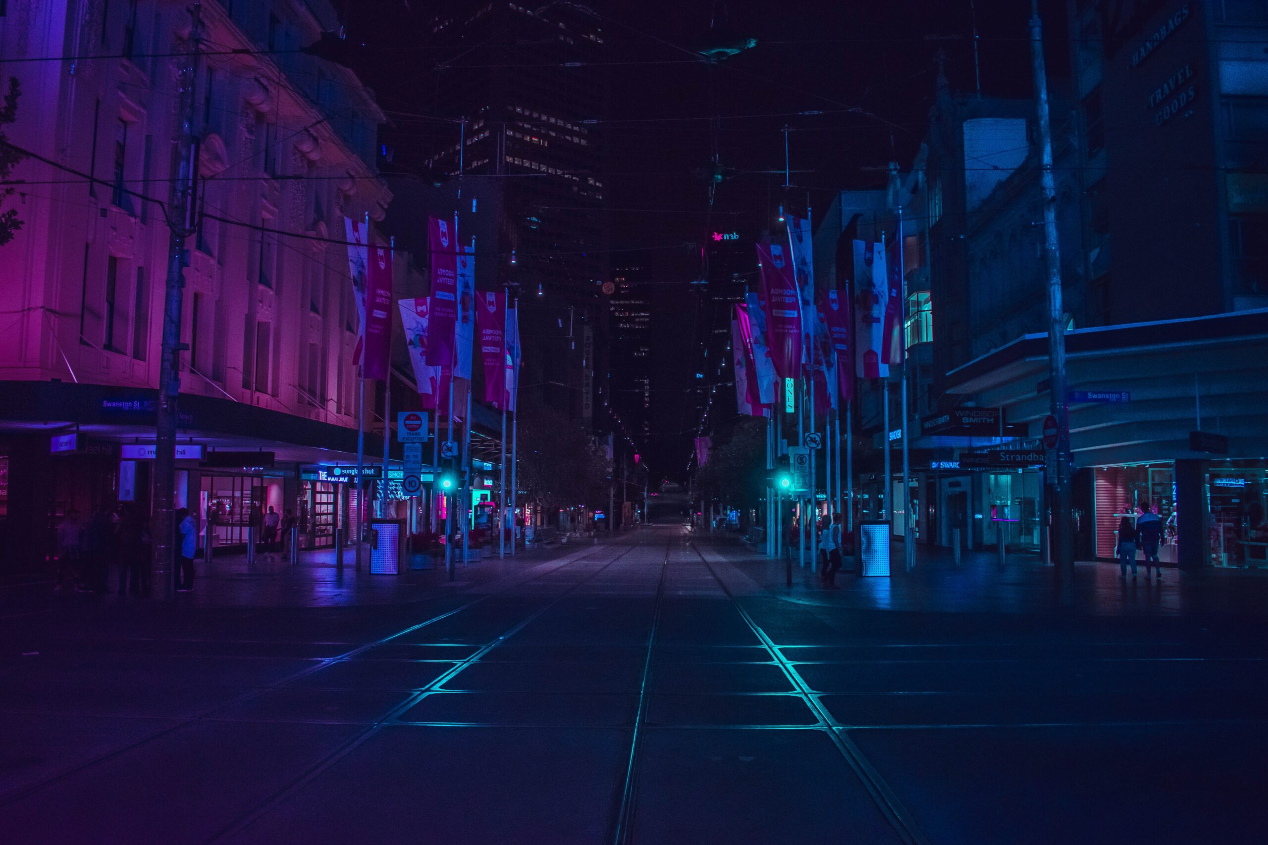 Der Blick bei Nacht auf eine Straße und umliegende Gebäude. Alles ist in bläulichen und Romanen Farben ausgeleuchtet, die Szene wirkt futuristisch.